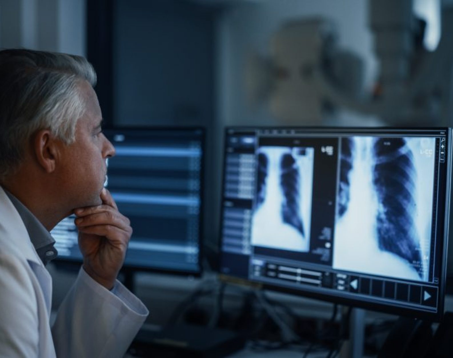 Experienced Radiographer Examines Chest X-rays on Multiple Computer Monitors in a Dark Medical Room. Modern Diagnostic Equipment Ready to Take Medical Imaging Scans of a Male Patient